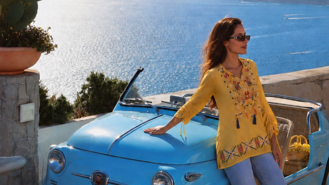 Woman in a yellow shirt standing next to a blue vintage car with a scenic ocean view in the background.